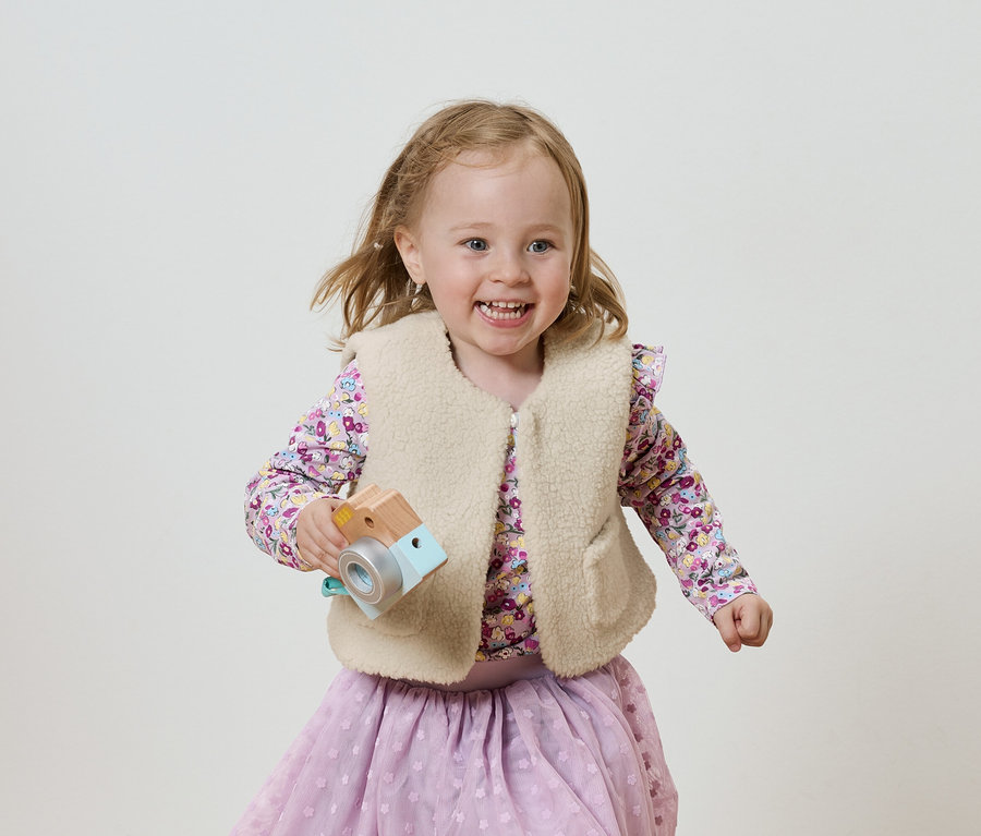 Fillette souriante avec un appareil photo en bois, vêtue d'un chemisier et d'une jupe lilas, sur un fond clair.