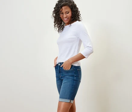 Une femme aux cheveux bouclés et foncés sourit et pose en bermuda en jean coupe « Lea », denim bleu moyen et un t-shirt blanc.