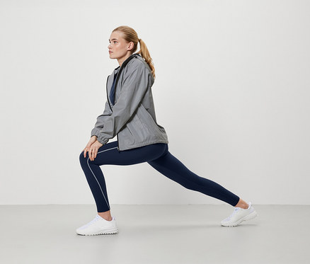 Femme blonde en veste grise et leggings bleus s'étire.