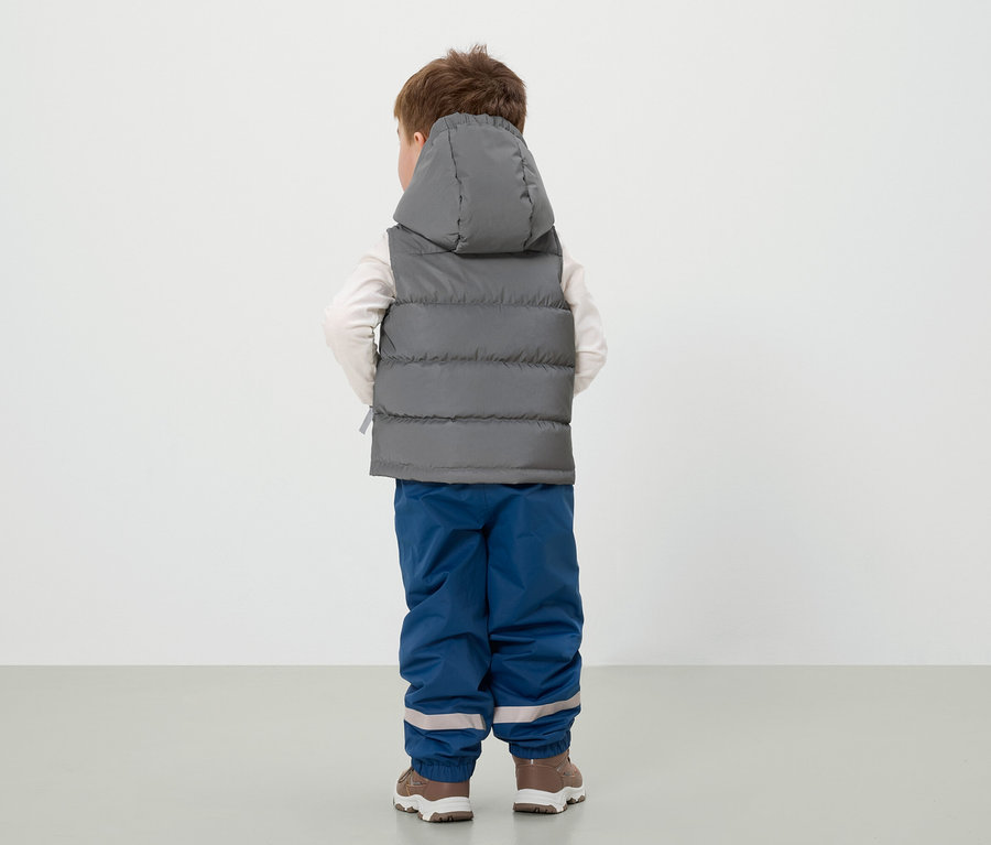 Un garçon, vu de dos, porte un gilet gris à capuche, un pantalon bleu et des chaussures marron.