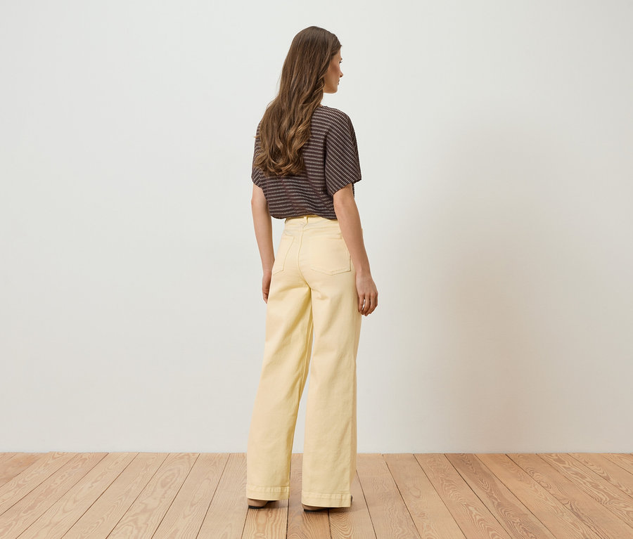 Vue de dos d'une femme portant un haut rayé marron et un pantalon jaune ample sur un plancher en bois devant un mur blanc.