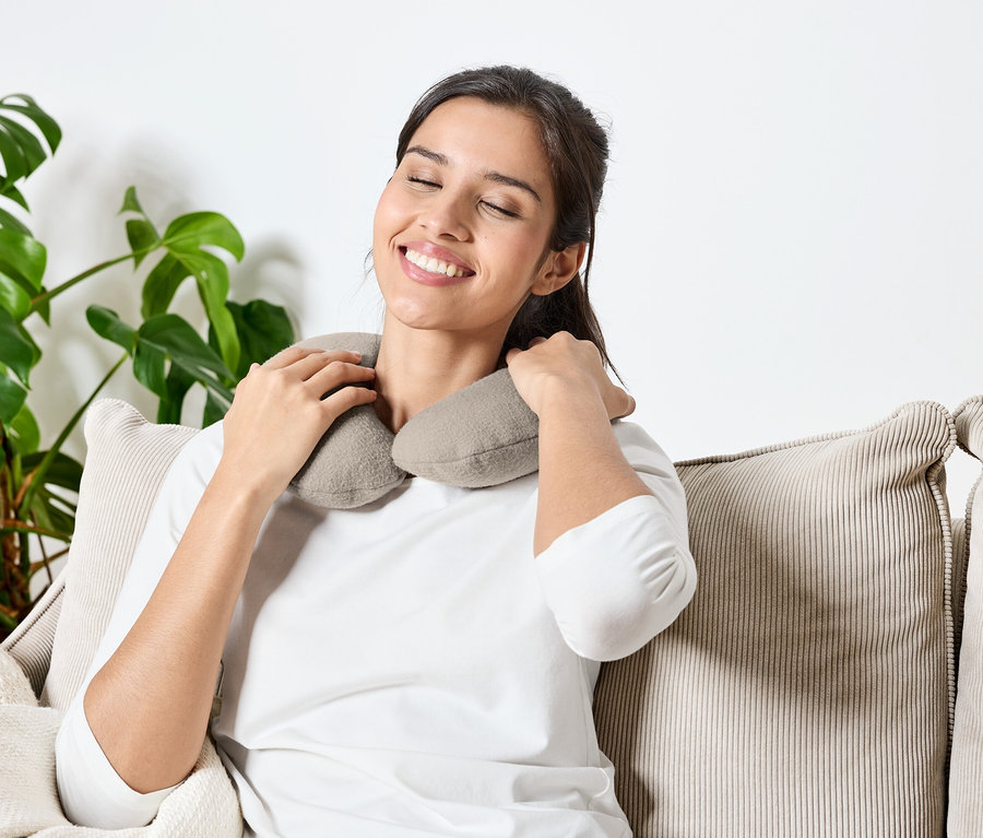 Une femme est assise les yeux fermés sur un canapé avec un coussin cervical gris autour du cou.