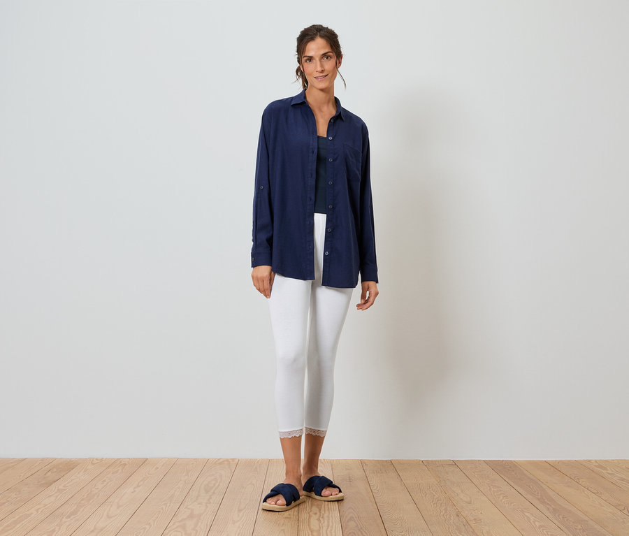 Femme debout devant un mur blanc portant une chemise bleue, un legging blanc et des pantoufles bleues.