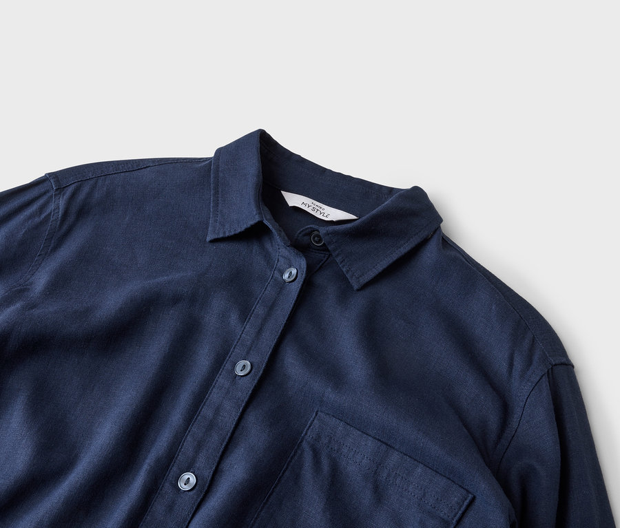 Gros plan d'une chemise bleu marine avec col, boutons et une seule poche poitrine sur un fond neutre.