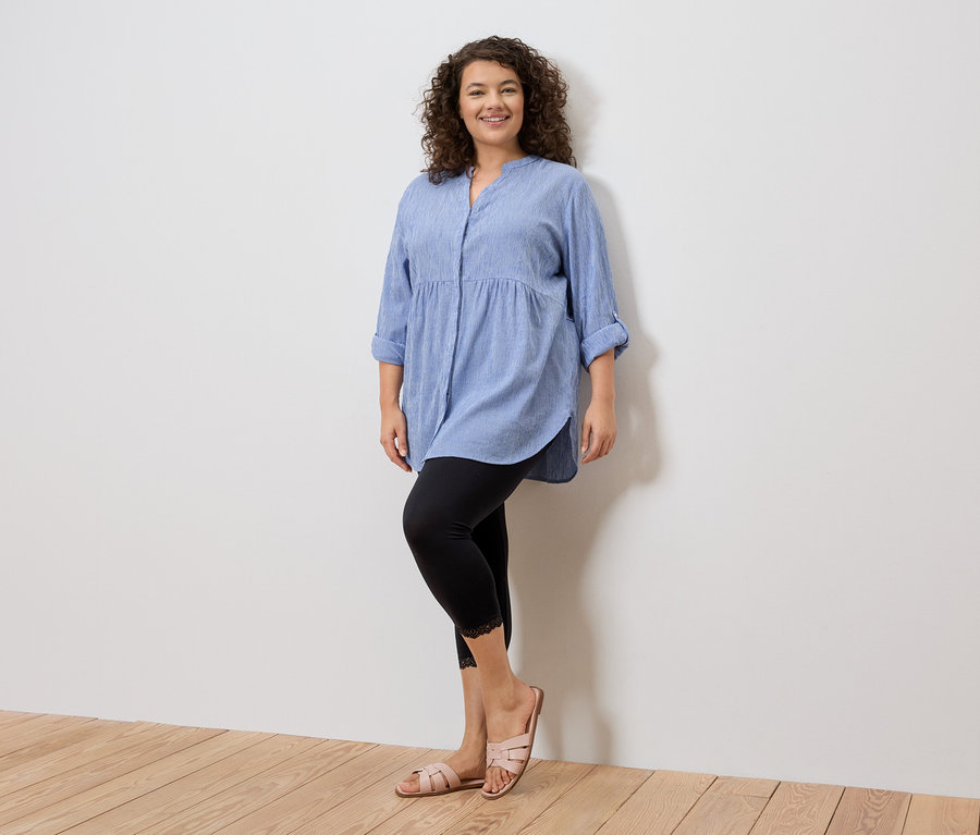 Femme debout devant un mur blanc portant une chemise bleue, un legging noir avec de la dentelle et des sandales roses.