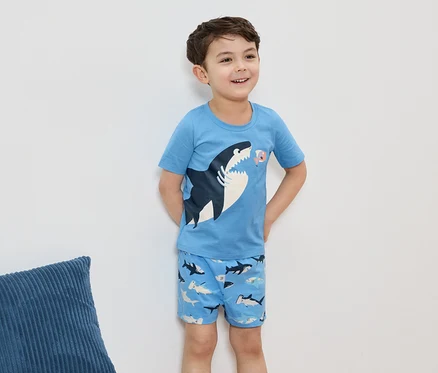 Un garçon souriant porte un pyjama bleu clair avec un motif de requin, devant un mur blanc.