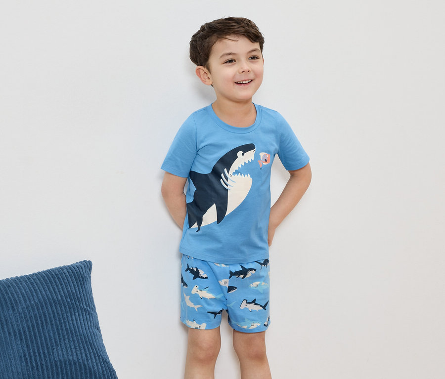 Un garçon souriant porte un pyjama bleu clair avec un motif de requin, devant un mur blanc.