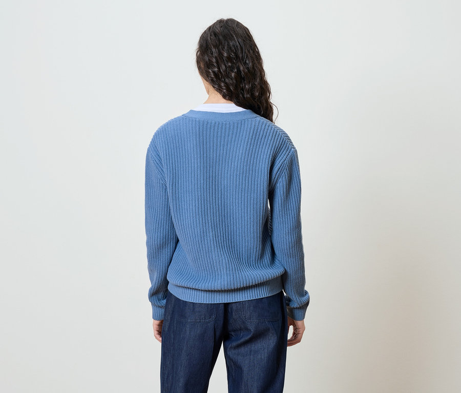 Une femme aux longs cheveux ondulés et foncés se tient dos à la caméra, portant un pull en tricot bleu et un jean.