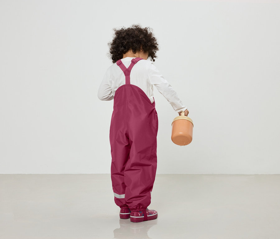 Enfant aux cheveux bouclés vu de dos, portant un T-shirt blanc à manches longues, une salopette bordeaux et des bottes en caoutchouc, tenant un seau beige.