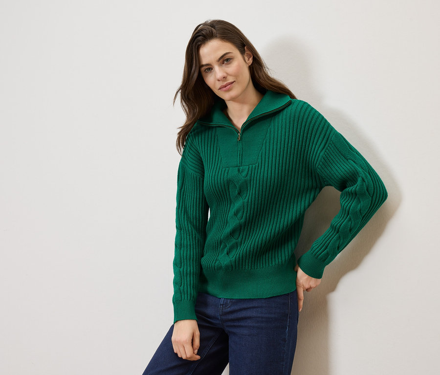 Une femme aux cheveux bruns pose devant un mur blanc, portant un pull en maille vert zippé et un jean bleu.