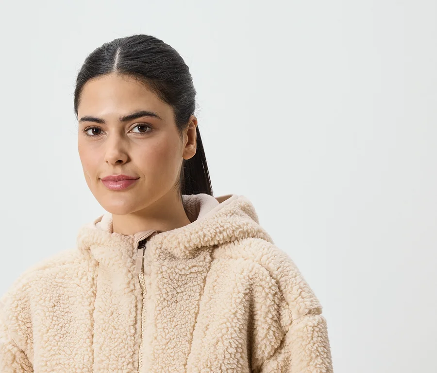 Une femme avec une queue de cheval porte une veste à capuche beige en tissu peluche sur fond blanc.