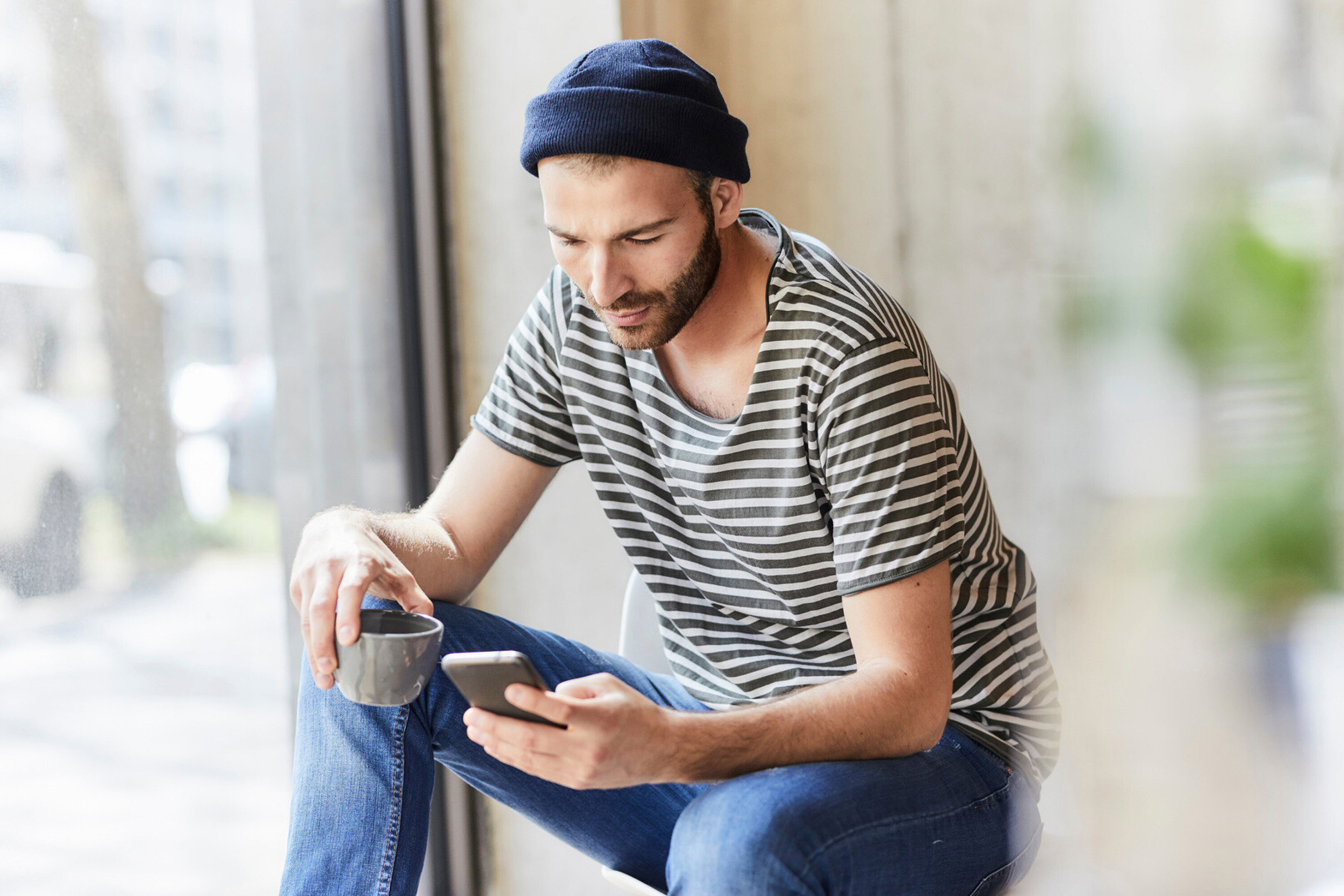 Un homme avec un bonnet et un t-shirt rayé est assis avec un téléphone et une tasse à la main.