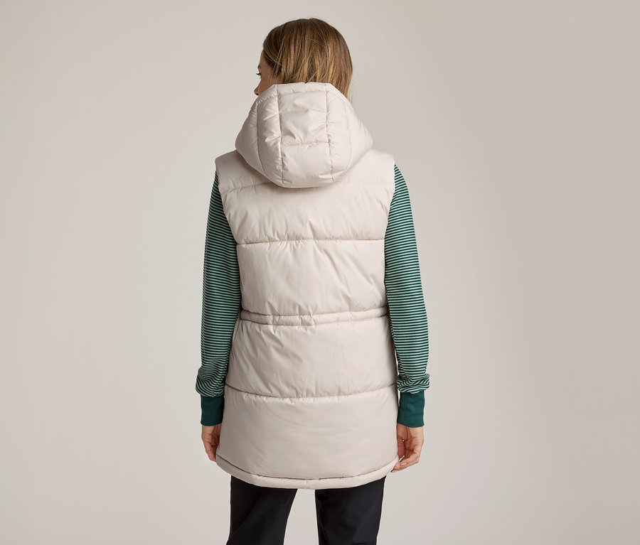 Une femme vue de dos, portant un gilet matelassé beige à capuche par-dessus un t-shirt à manches longues rayé vert et blanc et un pantalon foncé.