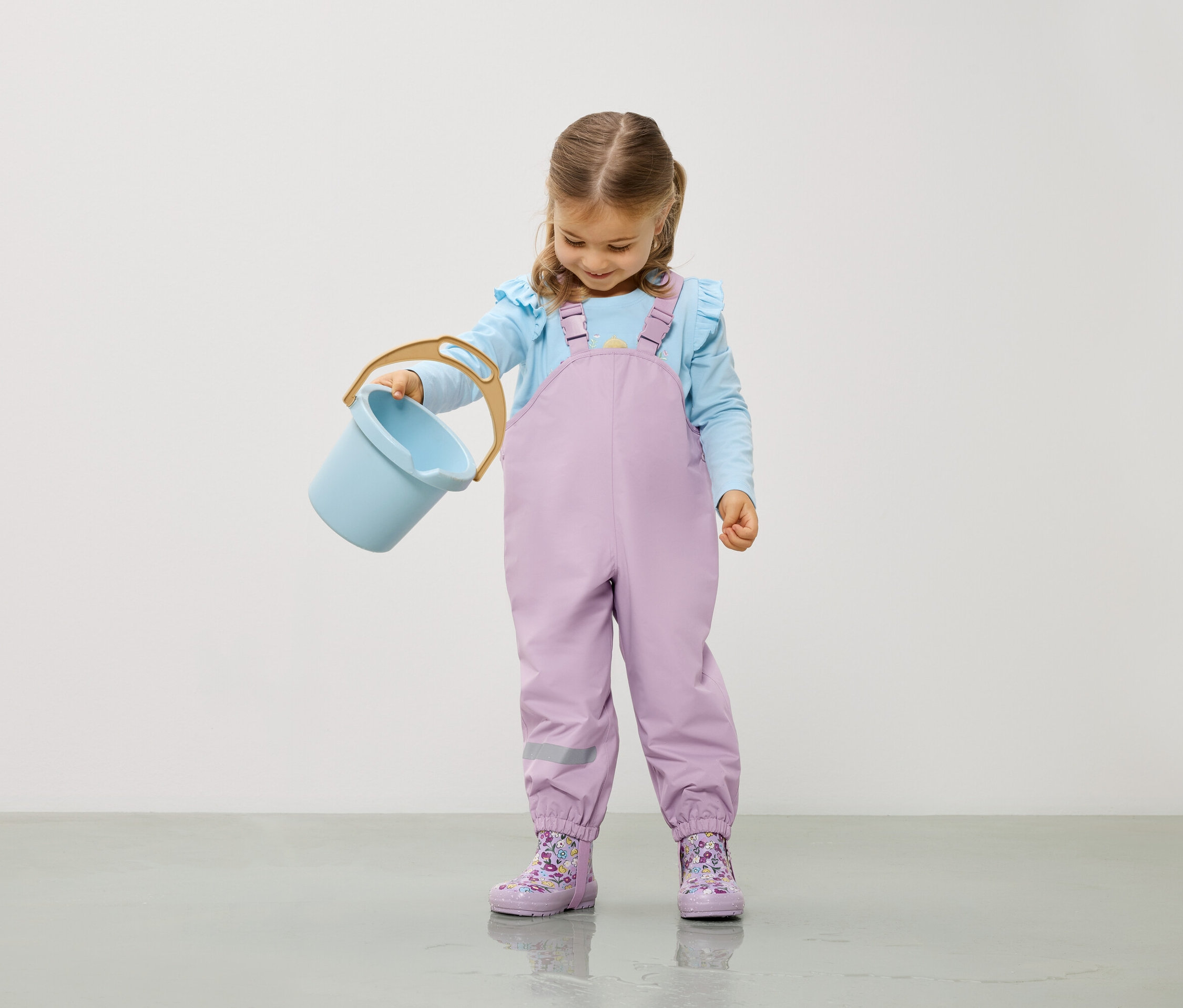 Fillette en chemisier bleu clair et salopette violette tenant un seau bleu clair.