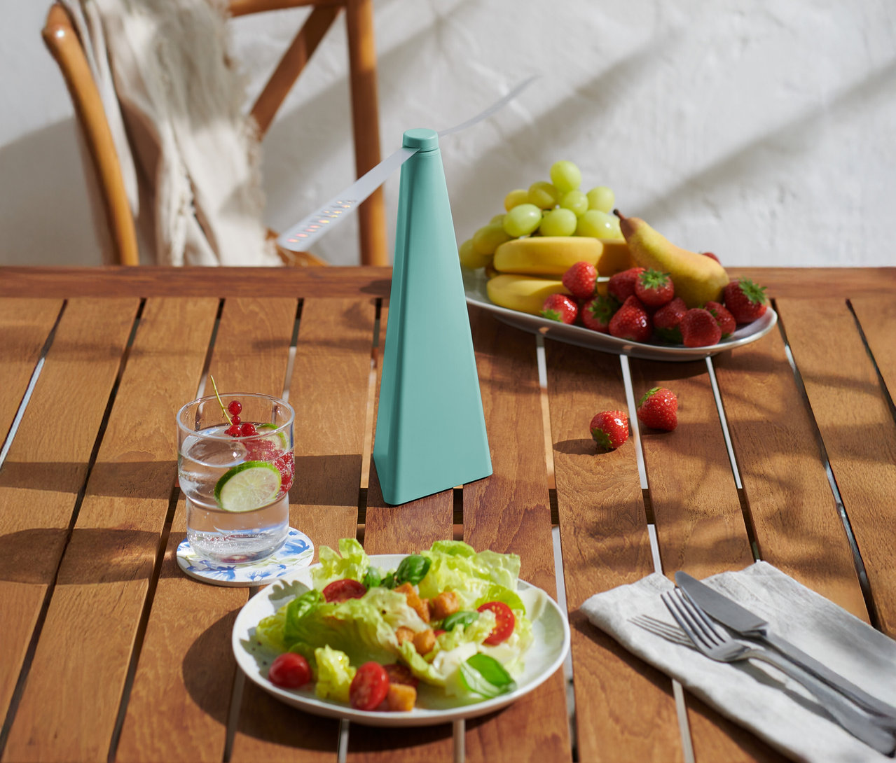 Sur une table en bois se trouvent une boisson, une assiette de salade, un plat de fruits et un ventilateur turquoise.