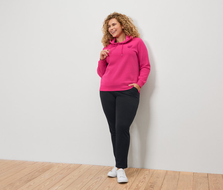 Femme aux cheveux bouclés portant un sweat à capuche rose et un pantalon noir.