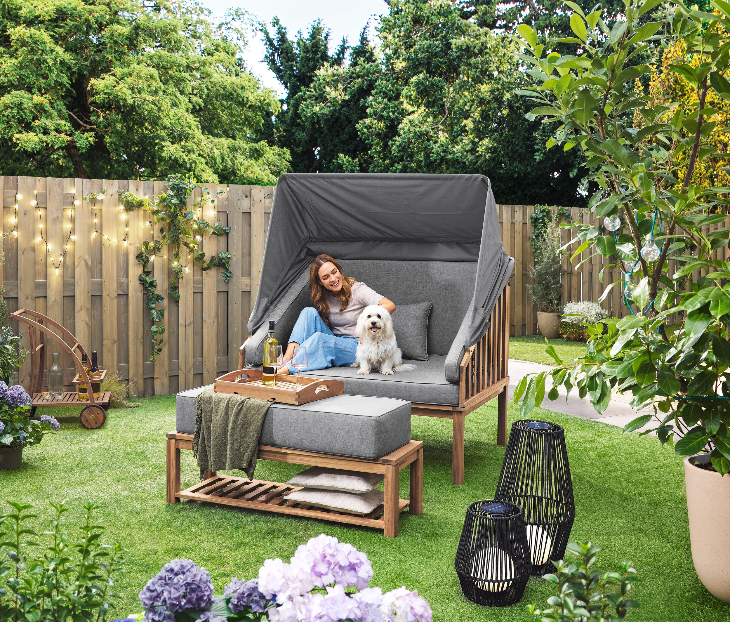 Une femme est assise avec un chien dans un Premium Lounge-Strandkorb mit Sonnenschutz-Dach. Une Lanterne solaire avec ruban en tissu, grand modèle et une Lanterne solaire avec ruban en tissu, petit modèle sont posées sur la pelouse. Un Barwagen mit abnehmbarem Tablett est visible à l'arrière-plan. Une LED-Outdoor-Lichterkette mit Netzadapter est accrochée à une clôture.
