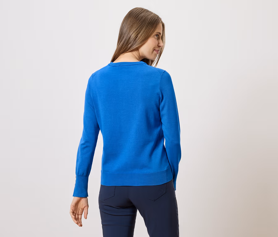 Une femme aux longs cheveux bruns, vue de dos, porte un pull bleu et un pantalon bleu marine sur un fond blanc.