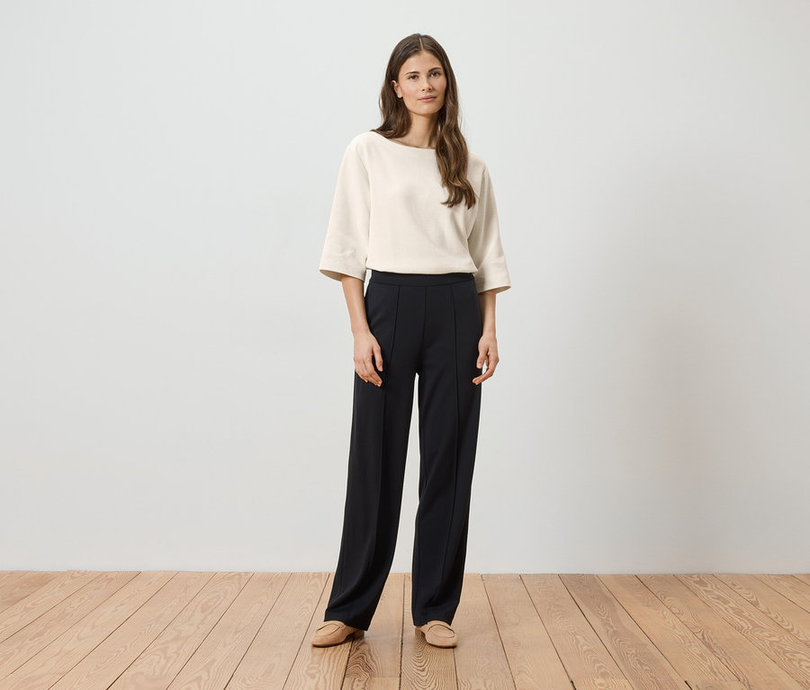 Femme portant un chemisier crème à manches trois-quarts et un pantalon noir à plis, avec des chaussures beiges.