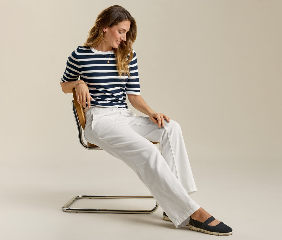 Une femme assise sur une chaise porte un jeans grand confort, un t-shirt rayé et des chaussures plates.