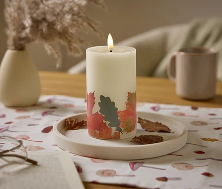 Une bougie blanche allumée avec un motif de feuilles est posée sur une table avec un pichet beige et un vase.