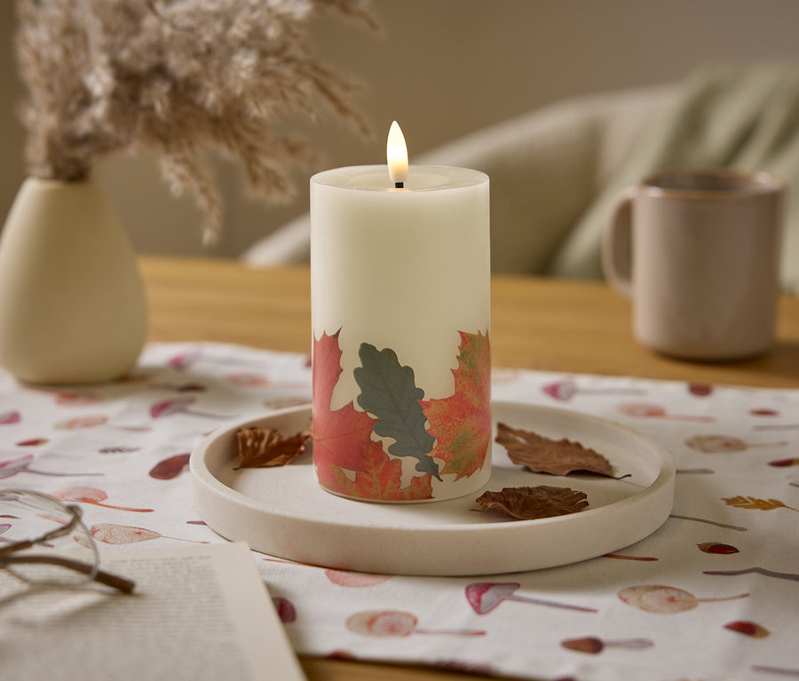 Une bougie blanche allumée avec un motif de feuilles est posée sur une table avec un pichet beige et un vase.