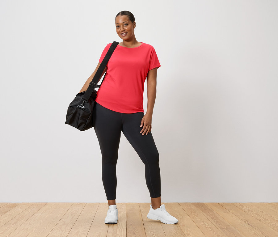 Femme portant un t-shirt rouge et des leggings avec un sac de sport noir sur l'épaule et des baskets blanches.