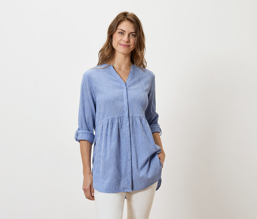 Une femme porte un chemisier bleu clair à manches longues et un pantalon blanc.