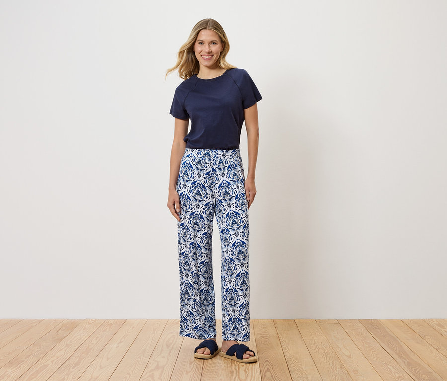 Une femme se tient devant un mur blanc, portant un t-shirt bleu marine, un pantalon bleu et blanc à motifs et des sandales bleues.