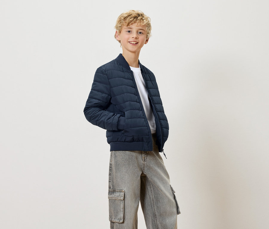 Un adolescent blond souriant porte une veste matelassée bleu marine et un pantalon cargo gris.