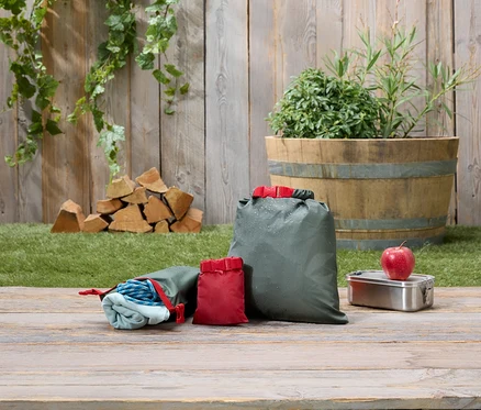 Trois sacs d'emballage olive et rouge sont posés sur une table en bois à côté d'une boîte à lunch et d'une pomme.