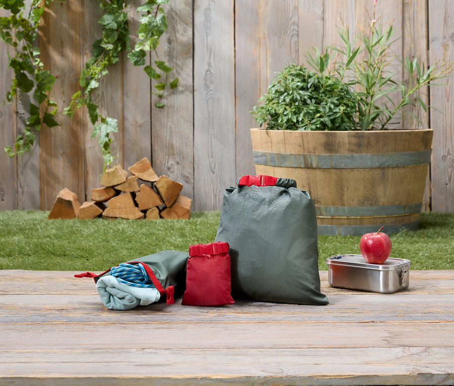 Trois sacs d'emballage olive et rouge sont posés sur une table en bois à côté d'une boîte à lunch et d'une pomme.