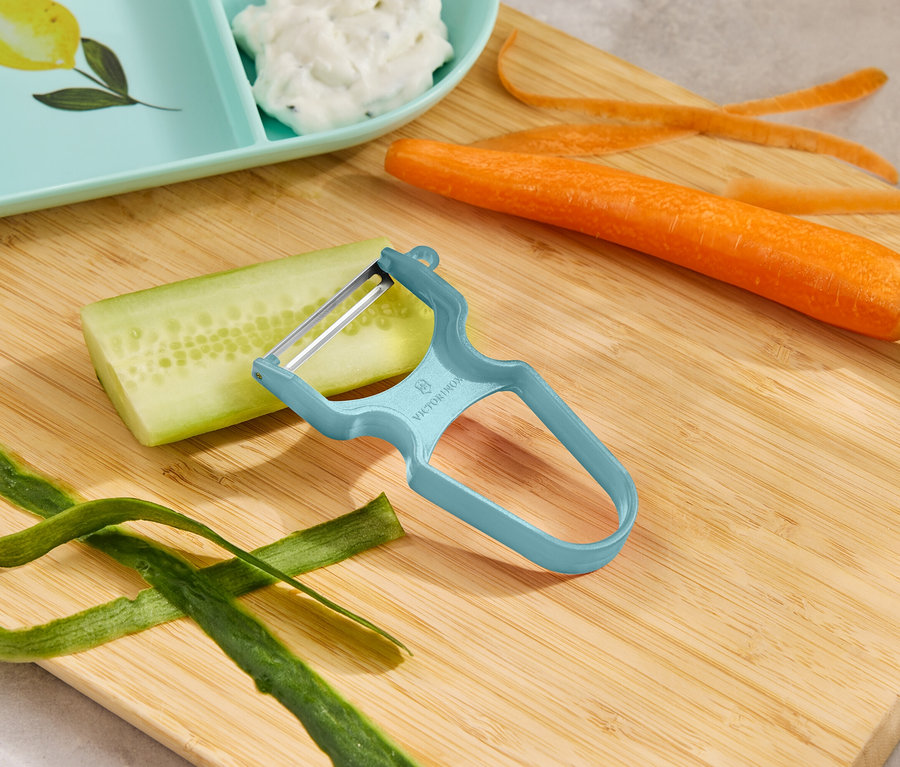 Un éplucheur à légumes bleu est posé sur une planche à découper en bois à côté d'un concombre, d'une carotte et de pelures de légumes.
