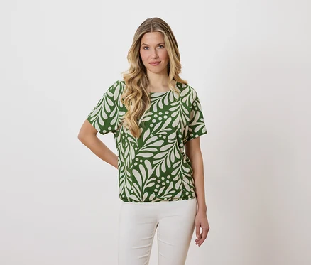 Femme aux cheveux blonds ondulés portant un chemisier vert à motifs de feuilles et un pantalon blanc.