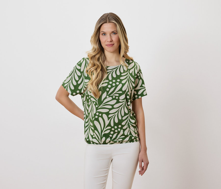Femme aux cheveux blonds ondulés portant un chemisier vert à motifs de feuilles et un pantalon blanc.