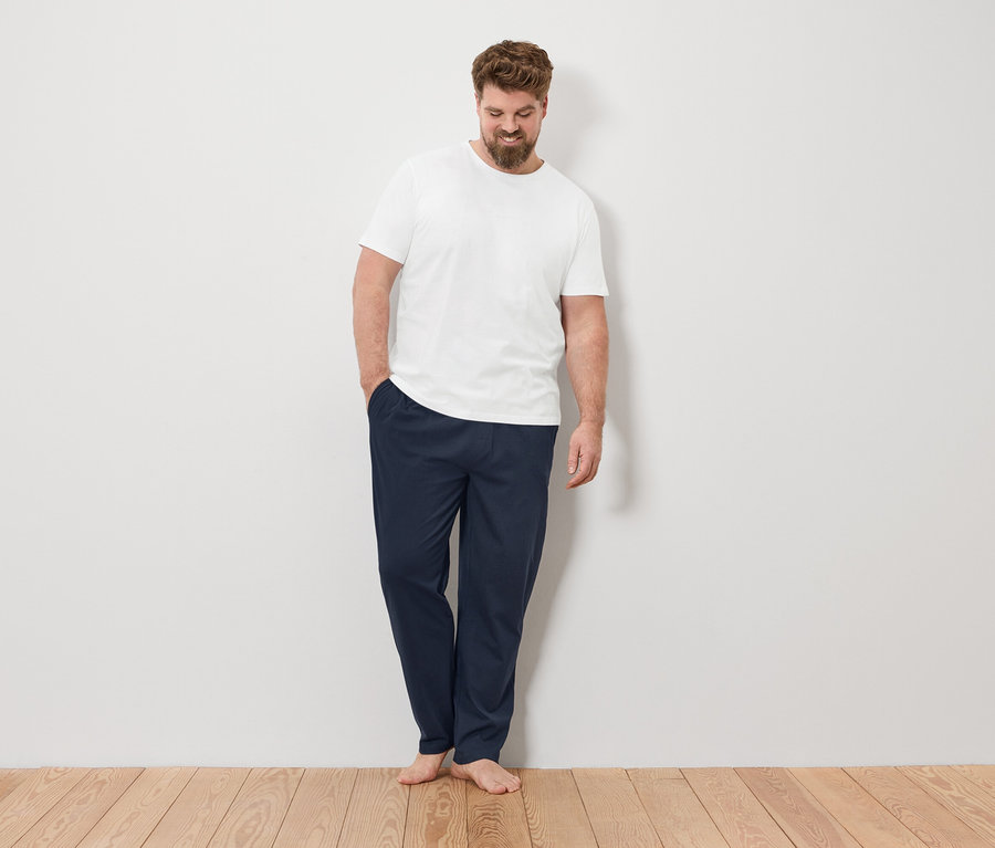 Un homme se tient pieds nus, la main dans la poche de son pantalon, devant un mur blanc.
