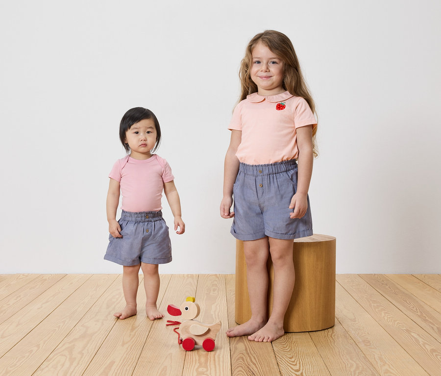Deux filles debout sur un plancher en bois, toutes deux vêtues de hauts roses et de shorts bleus. Un jouet en bois est sur le sol.