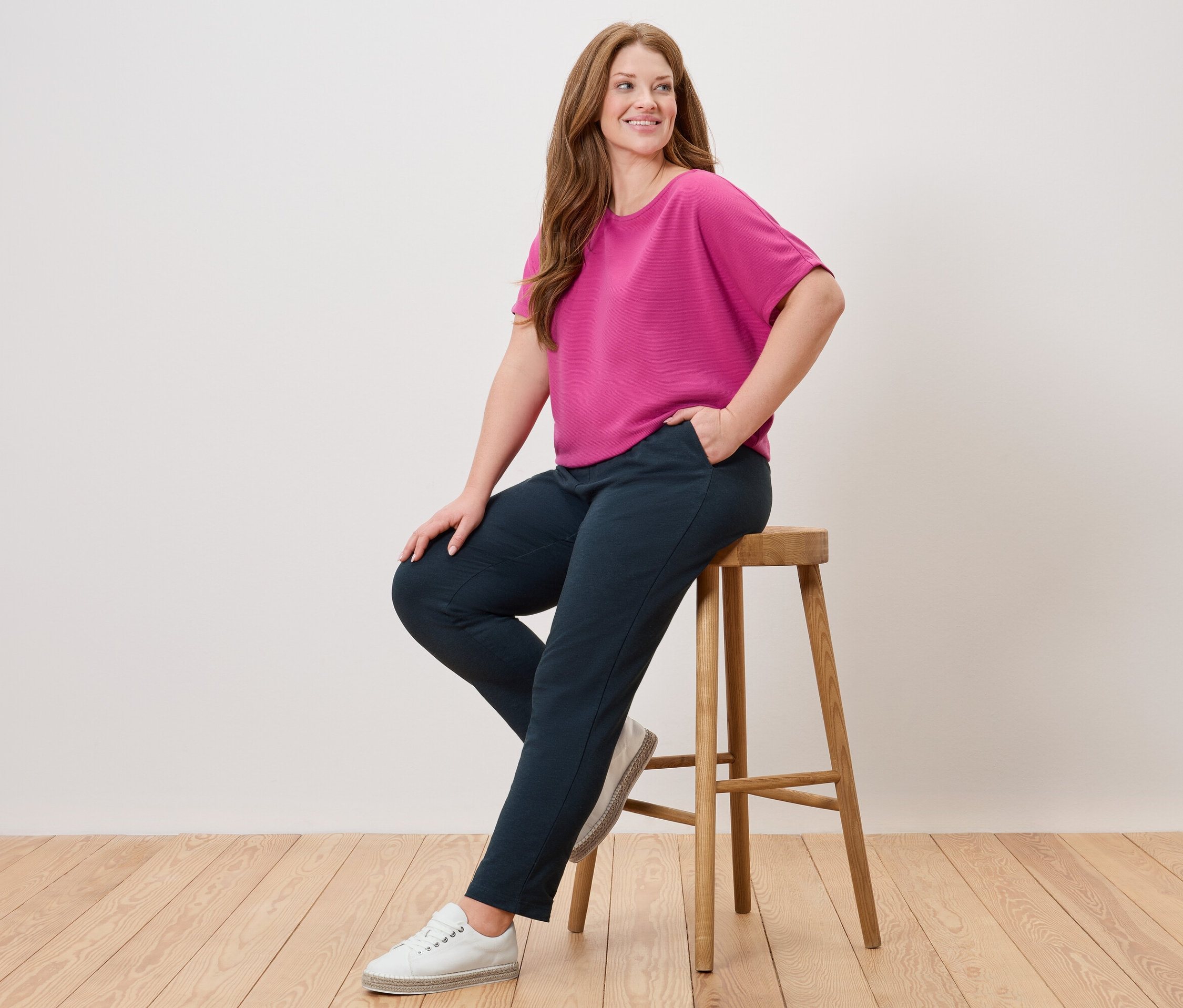 Une femme est assise sur un tabouret, portant un haut rose, un pantalon bleu et des chaussures blanches.