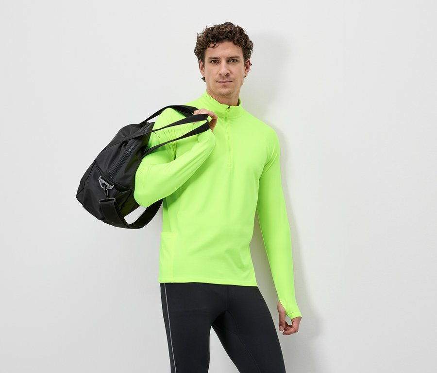 Un homme avec un sac de sport noir sur l'épaule, portant un t-shirt à manches longues jaune néon et un legging noir sur fond blanc.