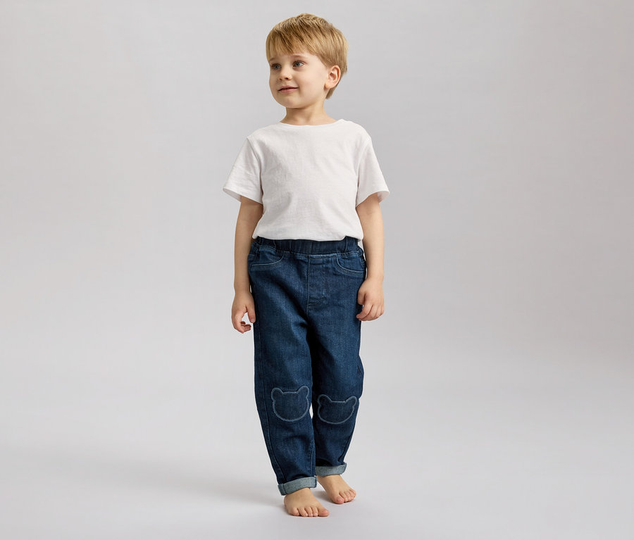 Un garçon blond se tient pieds nus sur un fond gris, vêtu d'un jean avec des motifs d'ours et d'un t-shirt blanc.
