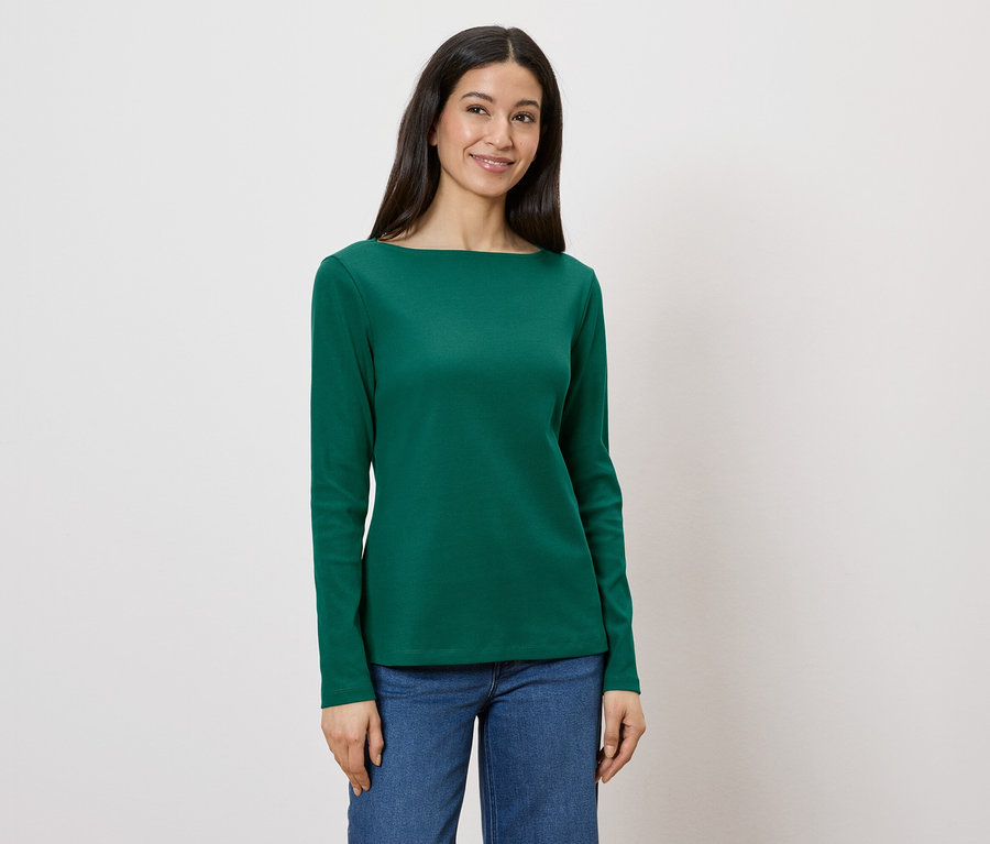 Une femme porte un chemisier vert à manches longues et un jean sur fond blanc.