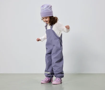 Une fillette aux cheveux bouclés porte un bonnet violet, un chemisier blanc, une salopette violette et des bottes roses.