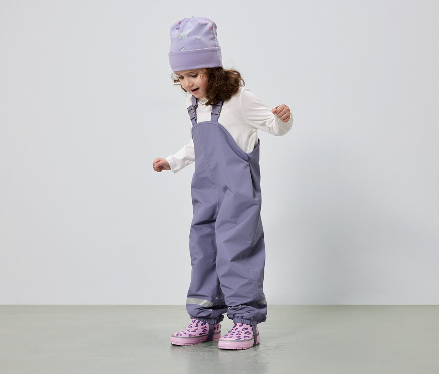 Une fillette aux cheveux bouclés porte un bonnet violet, un chemisier blanc, une salopette violette et des bottes roses.