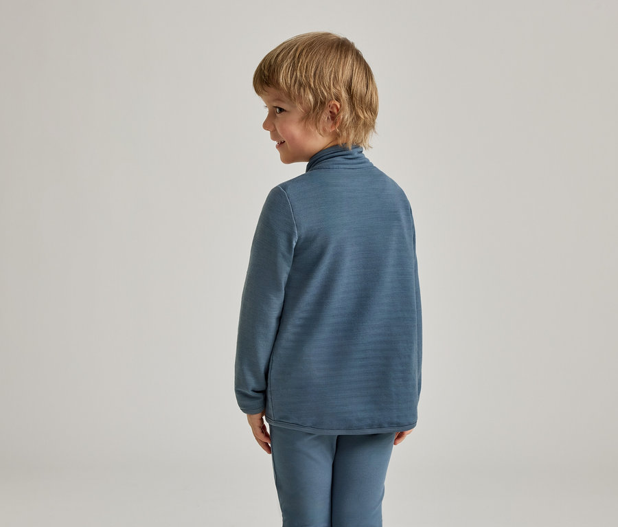 Un jeune enfant aux cheveux blonds, vu de dos et de profil, porte un haut bleu à manches longues avec un col montant et un pantalon bleu assorti. L'enfant regarde vers la droite avec un léger sourire.