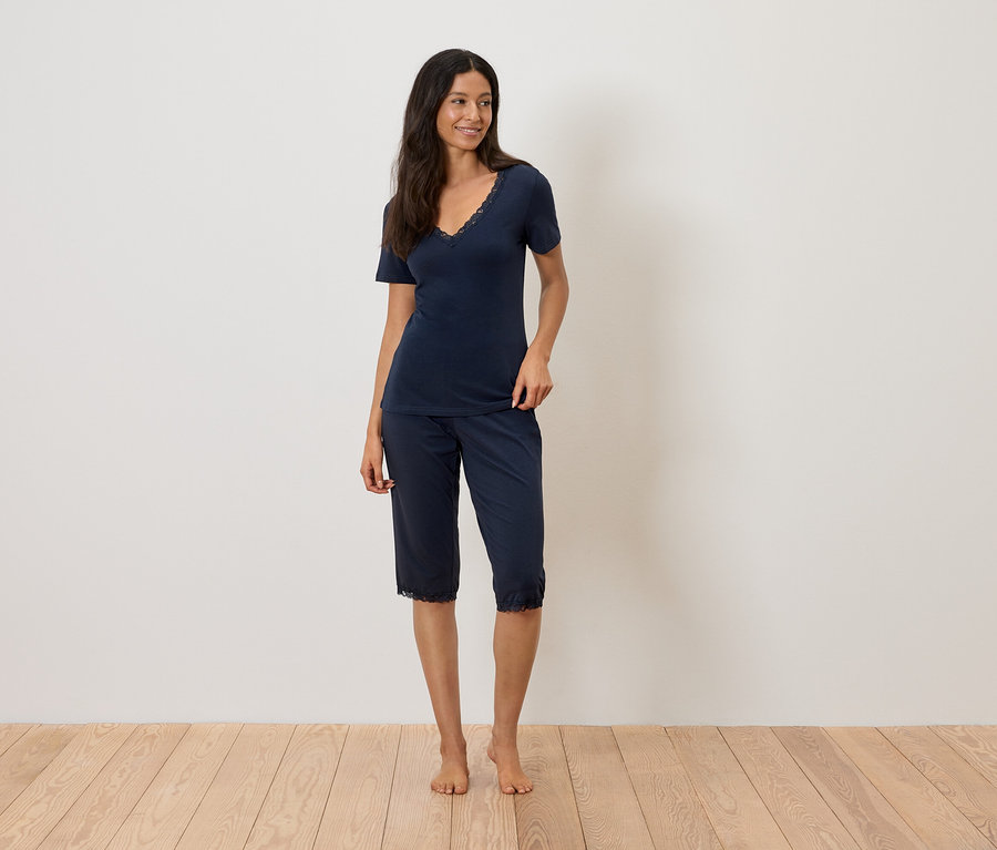 Une femme se tient pieds nus sur un plancher en bois, portant un pyjama bleu marine avec des détails en dentelle.