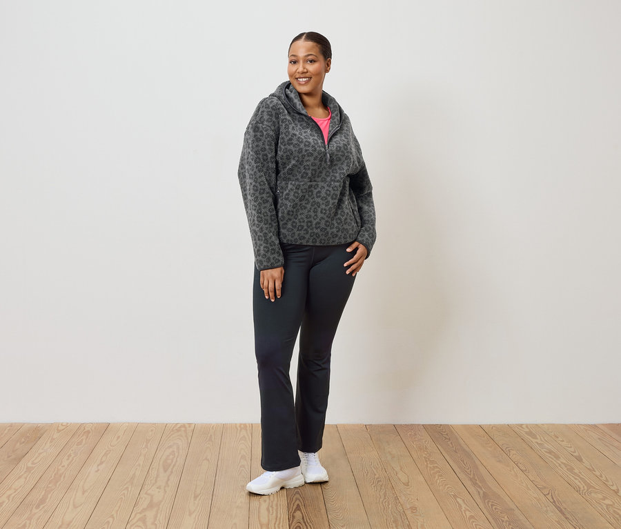 Une femme se tient debout et porte un sweat à capuche en polaire léopard gris et noir, un pantalon noir et des baskets blanches.