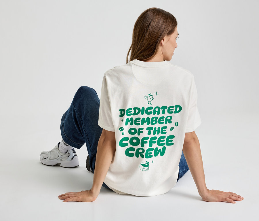 Femme assise par terre, portant un t-shirt avec l'inscription "Dedicated Member of the Coffee Crew", un jean et des baskets.