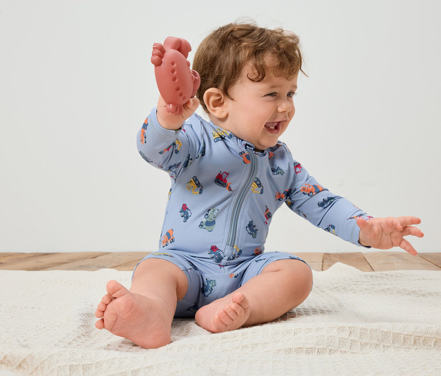 Un bébé souriant aux cheveux bouclés est assis sur une couverture blanche et tient un anneau de dentition rouge à la main. Le bébé porte une combinaison bleu clair avec des véhicules colorés.