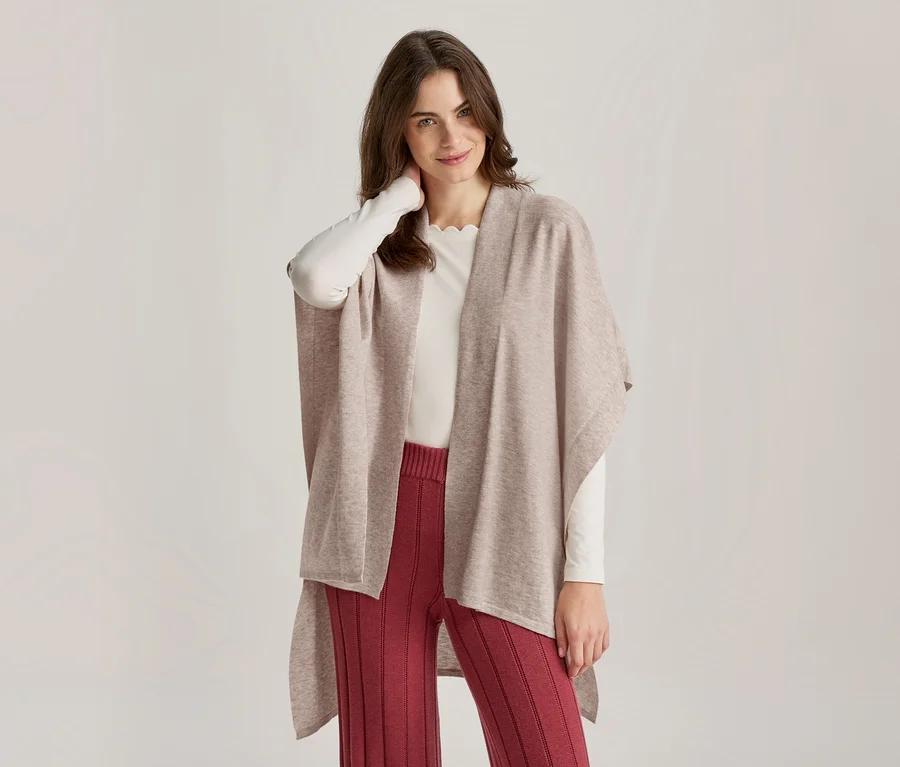 Une femme souriante portant un poncho beige, un haut blanc à manches longues et un pantalon côtelé rouge, une main derrière la nuque.