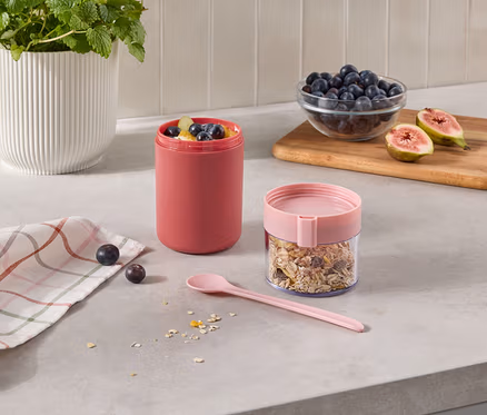 Deux récipients sur un comptoir de cuisine: un grand récipient rose rempli de fruits et un récipient transparent avec du muesli et un couvercle rose. À côté, il y a une cuillère rose, un torchon à carreaux, des baies, un bol de myrtilles et des figues coupées sur une planche en bois.